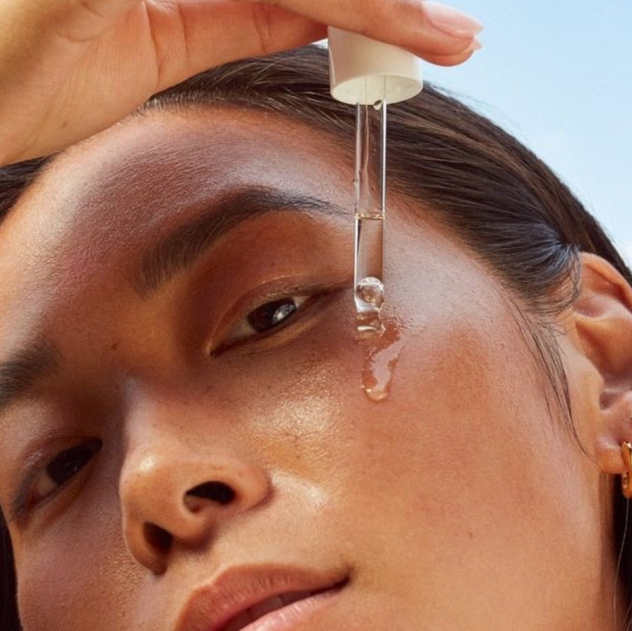 Person applying a dropper of liquid to their face against a clear sky.