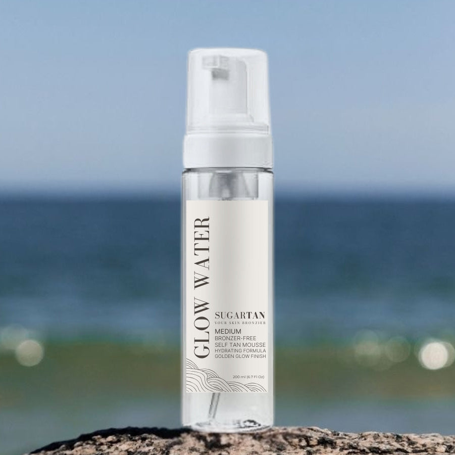 Skincare bottle labeled 'Glow Water' by Sugatan on a beach with ocean in the background